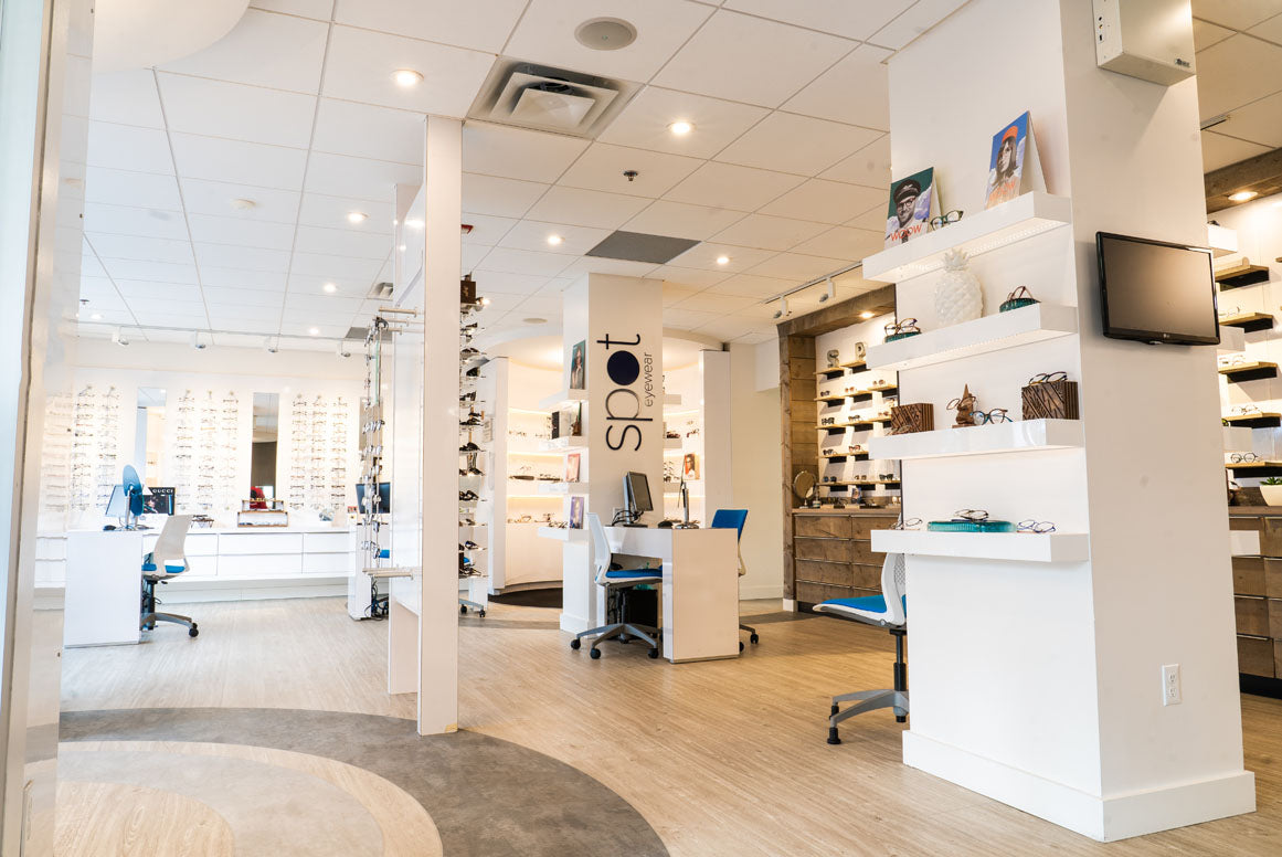 Wide view of modern optical shop. Hundreds of glasses on the walls and shelves. Bright, white room with beige wooden floors