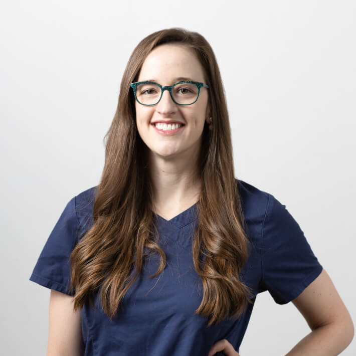 Dr Leanne Roach in blue scrubs. She's a young woman, long brown hair, blue glasses, smiling warmly.