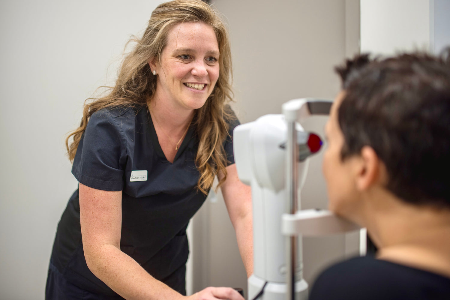 Eye technician in scrubs smiling and looking at a patient. She's controlling an instrument to measure the eye.