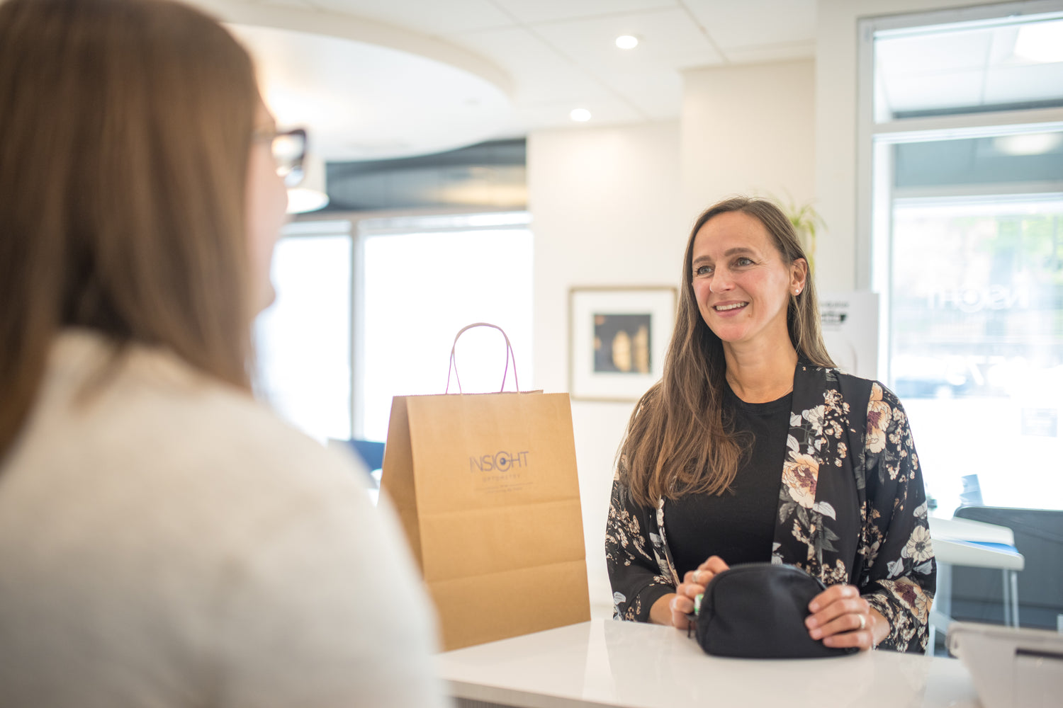 A female customer is being greeted by a reception team member after her appointment. A brown bag with the Insight Optometry logo is located beside the customer.