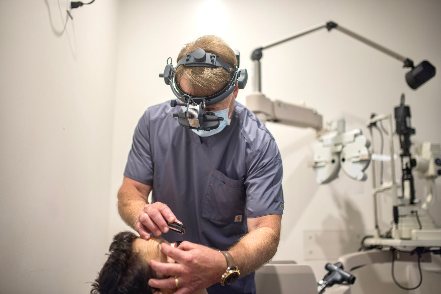 Woman in an eye exam chair, head leaning back. Male eye doctor wearing medical headgear leaned over her looking in her eye