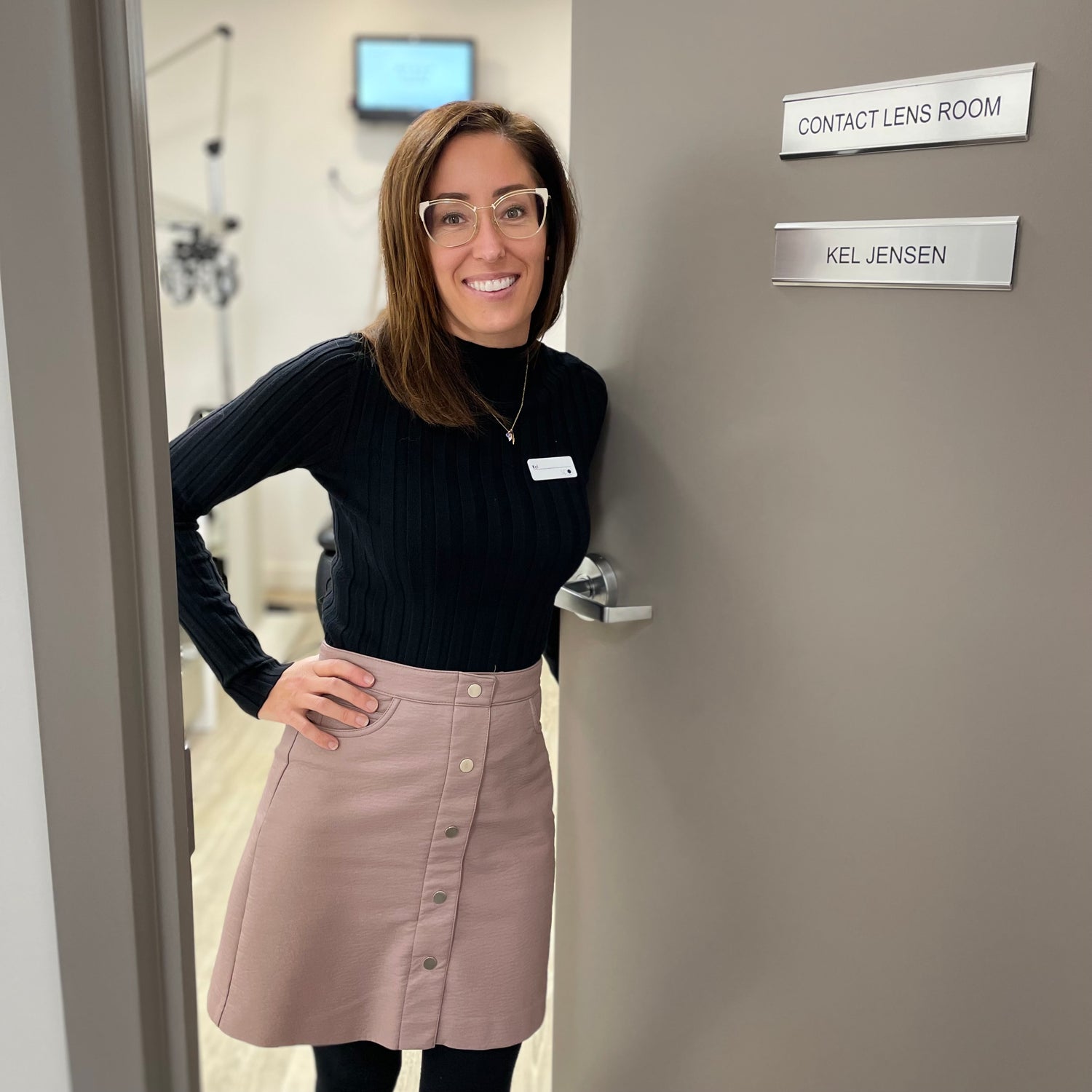 Friendly, smiling clinician standing at the doorway to an eye exam room. Door sign says Contact Lens Room, Kel Jensen.