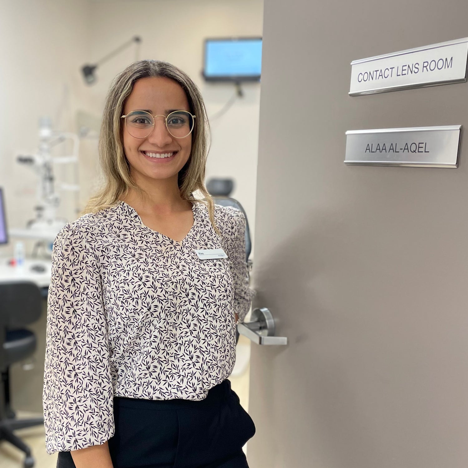 Smiling clinician welcoming you into an exam room.  The door sign says Contact Lens Room, Alaa Al-Aqel.