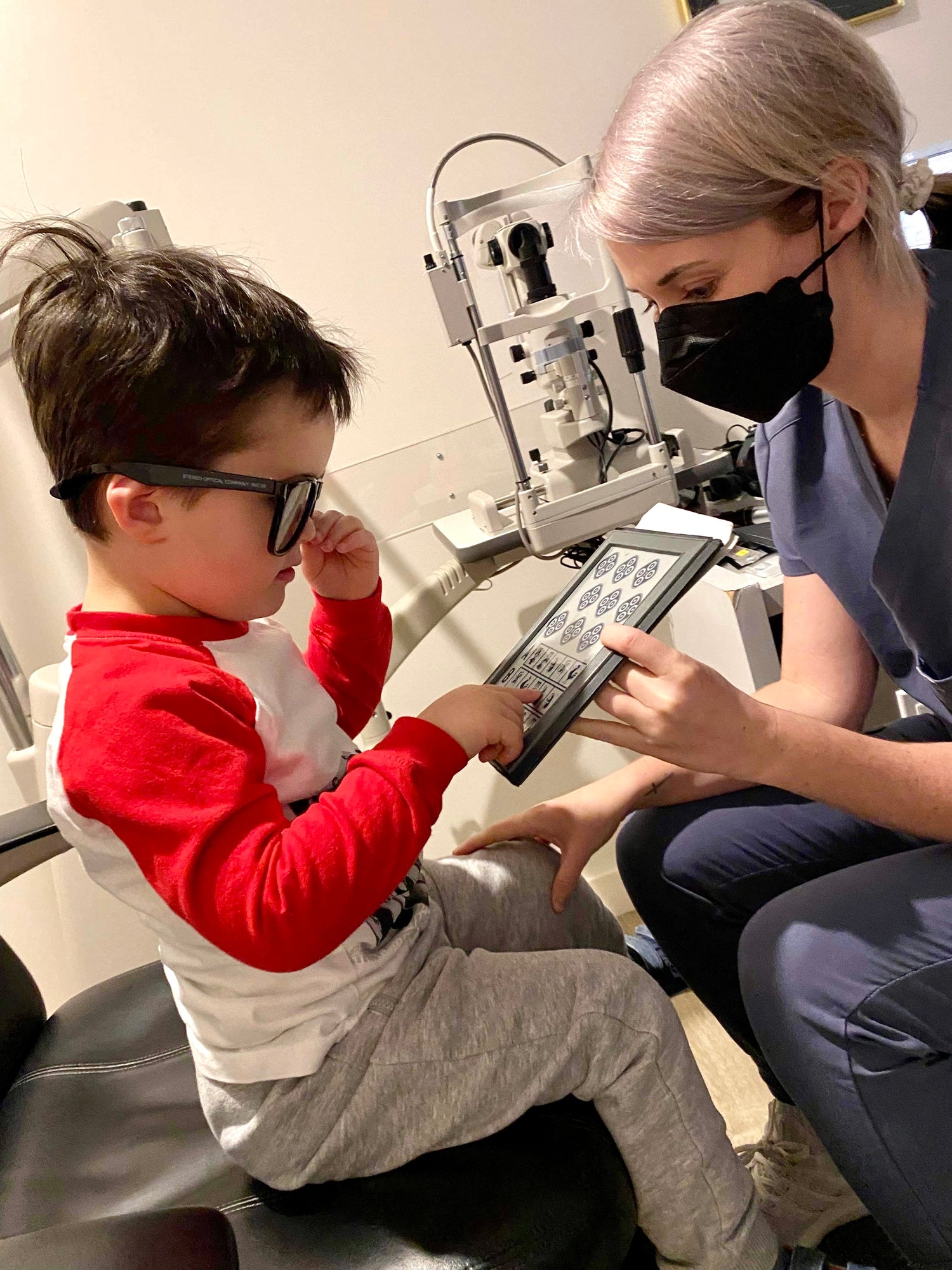 4 year old boy having eye exam, wearing glasses and pointing to pictures in a book that his eye doctor is holding.