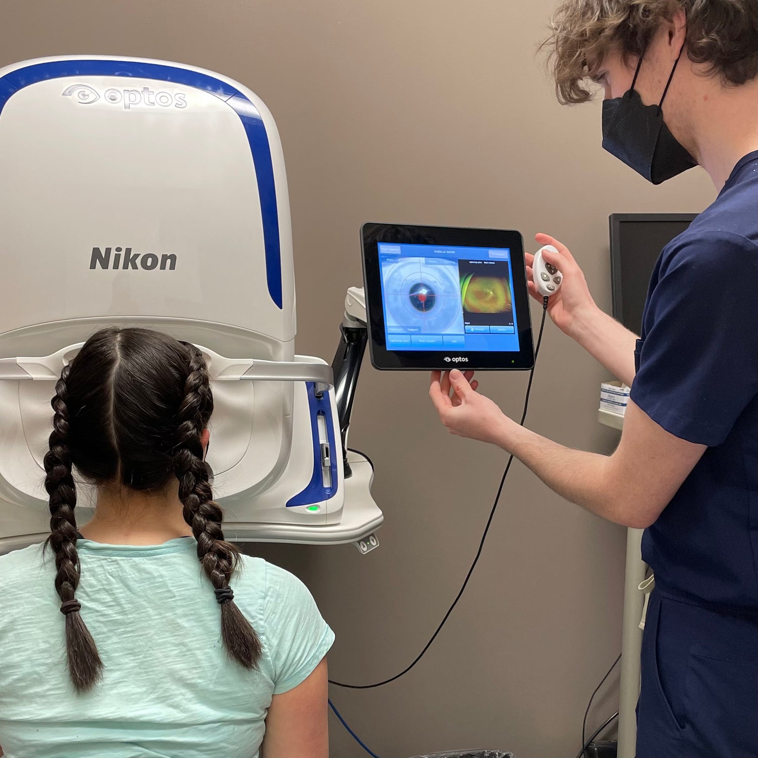 Child in braids sits in front of large intrument. Next to her a man in scrubs looks at a tablet, taking a picture of her eye