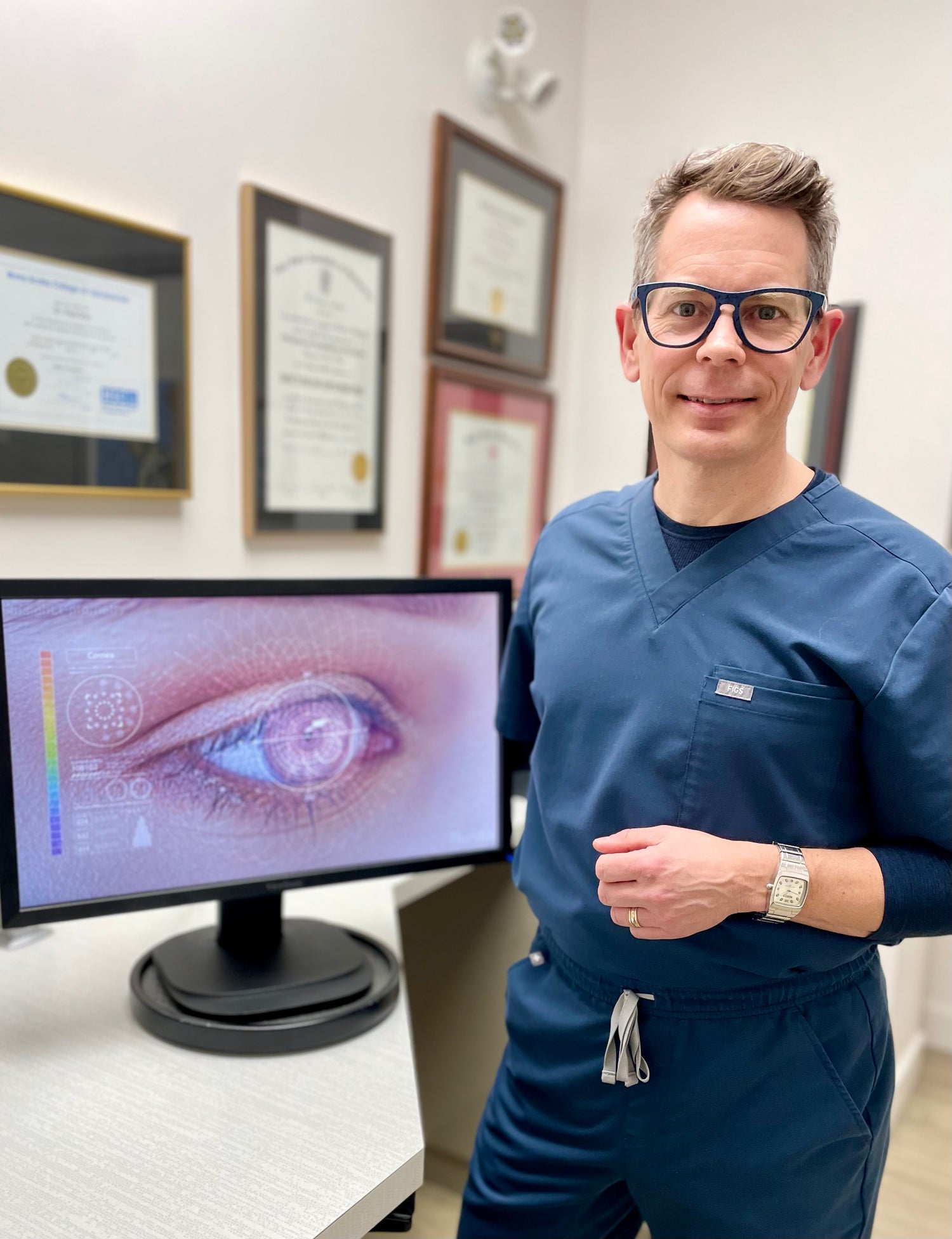 Smiling Dr Jeff Sansgter wearing scrubs, next to a computer screen with picture of an eye having laser surgery.
