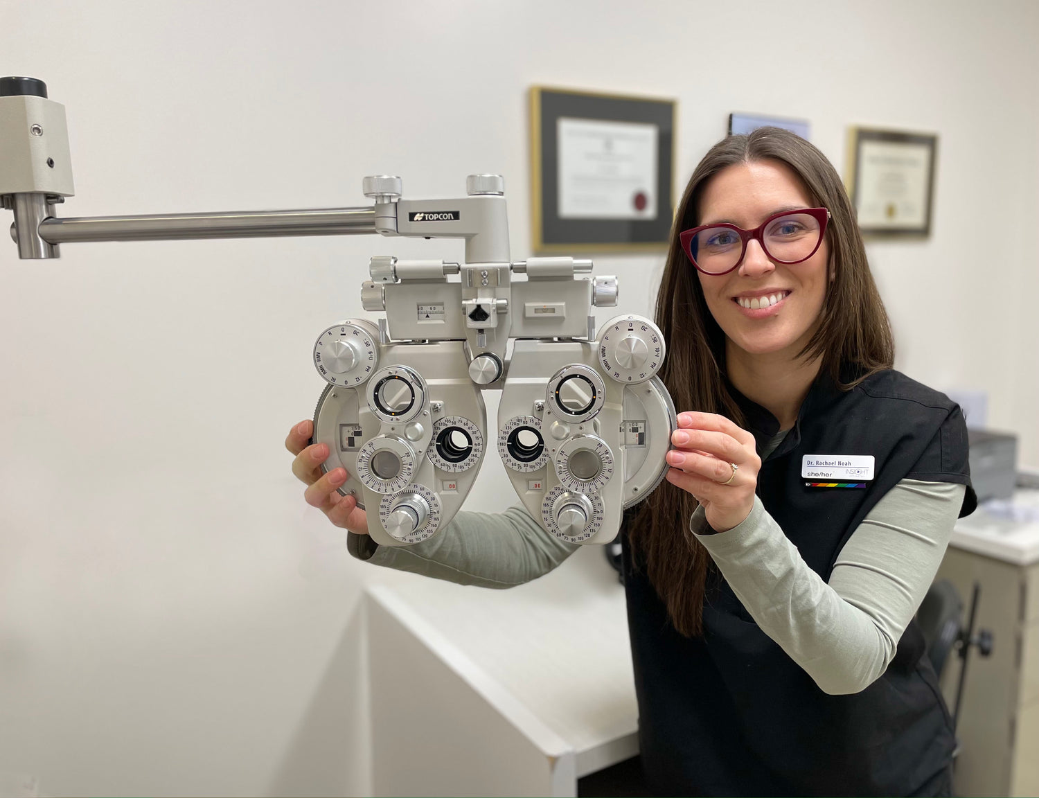 Dr Rachael Noah, smiling, in an eye exam room holding up an photropter instrument used to measure glasses prescriptions.