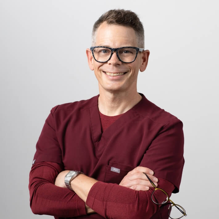 Dr Jeff Sangster in burgundy scrubs, arms folded, smiling energetically.