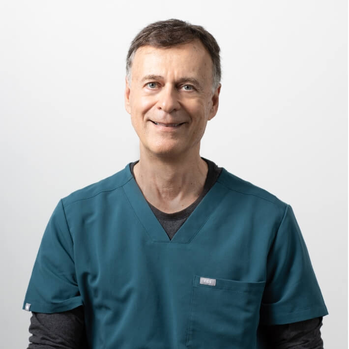 Dr Paul Gray wearing turquoise srubs. He's middle aged, has short dark hair and is smiling warmly.