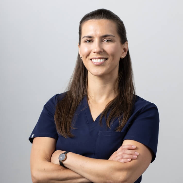 Dr Rachael Noah wearing scrubs. She's a young woman, long brown hair, arms folded, smiling warmly.