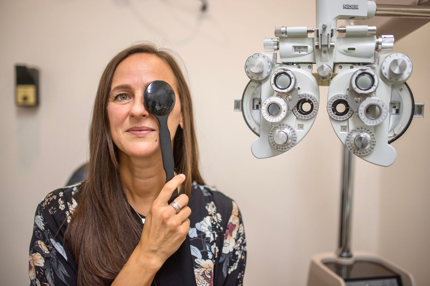 Long haired woman having eye exam, holding a small paddle over one eye to read chart. Next to her is optometry equipment.