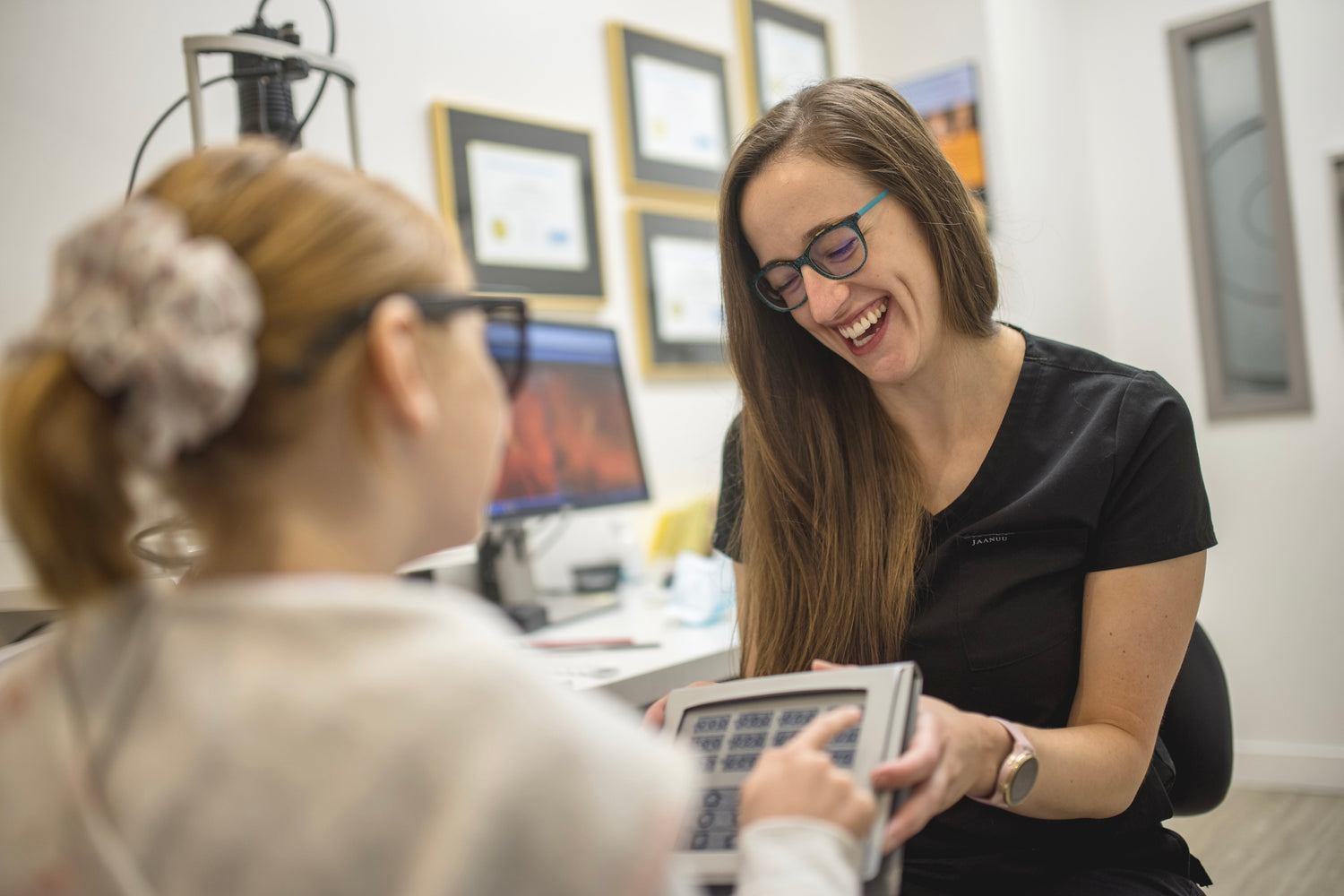 Dr. Leanne Roach is smiling while while holding a chart during an eye exam. A child, wearing special black glasses is pointing at the chart.